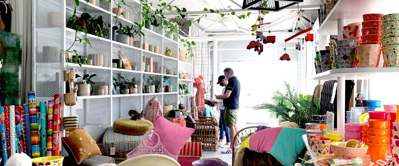 two people browse wares in light spacious store with shelves of vibrant cushions, plants, candles, wrapping paper and more