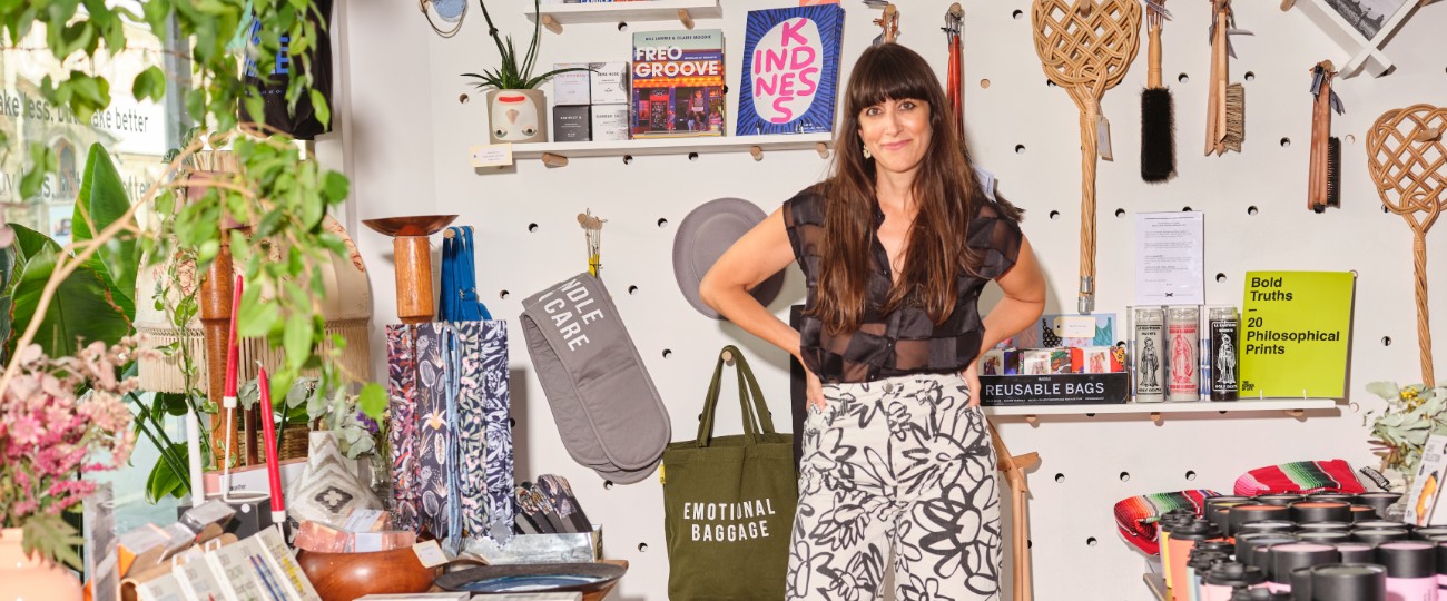 person with long dark hair, dark top and light patterned skirt stands in shop with colourful books, bags, plants and gifts