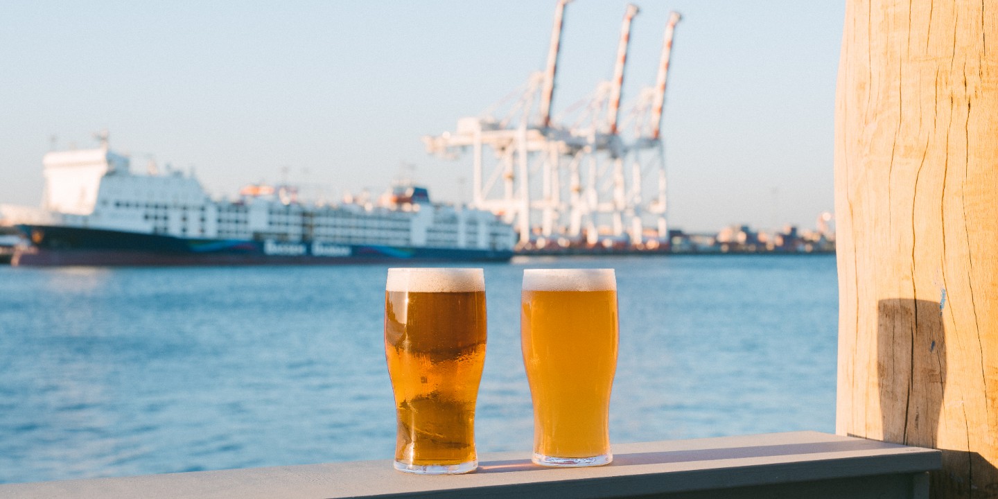 two cold pints rest in front of ocean, port cranes and big ship