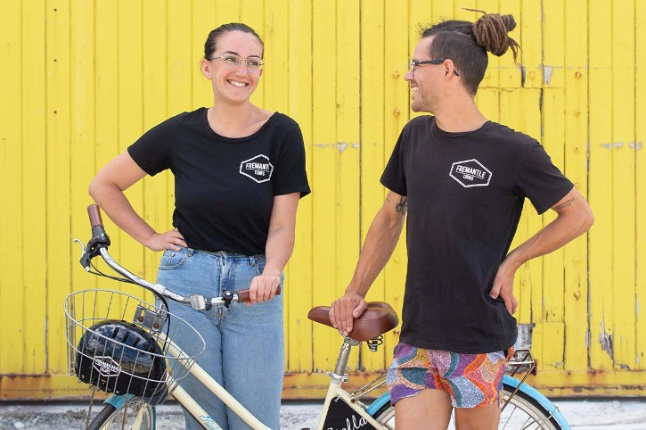 two smiling people wearing black t-shirts stand with bicycles against a bright yellow wall