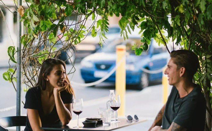 a couple chat easily to one another with glasses of red wine under lush greenery at Strange Company (Perth's best bars)