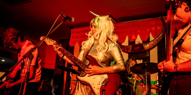 performer in glam costume and wig plays guitar with band on stage