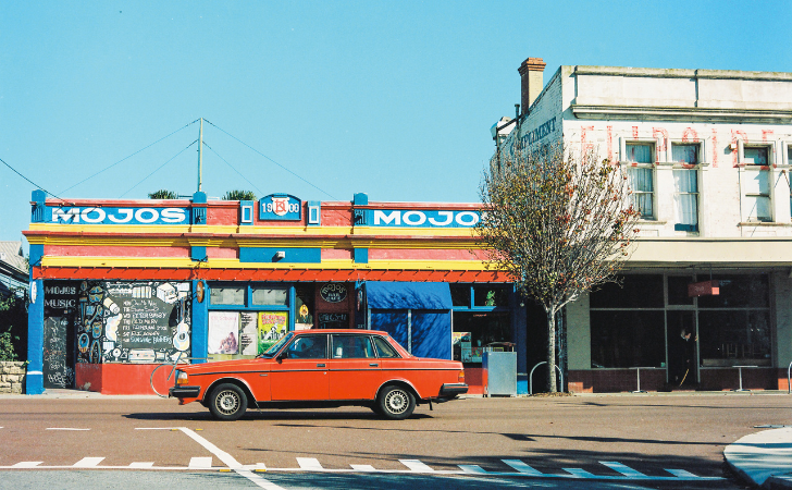 Red vintage car drives past iconic red, blue and yellow of Mojos in North Fremantle