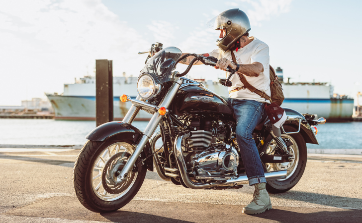 Jack Stillman has his leather bag across his body as he sits on a cafe racer-style motorbike in front of an ocean liner in Fremantle