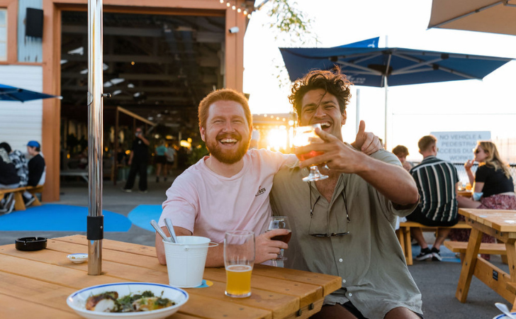 Two dads smiling with beers as the sun sets over the water in Fremantle at Gage Roads Freo