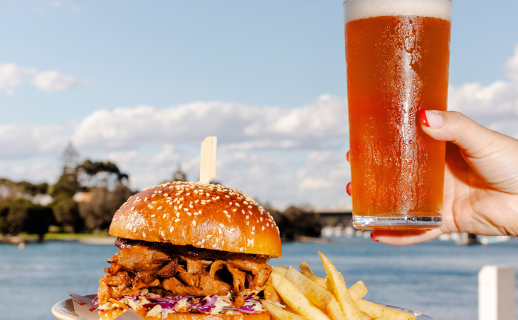 A hand holds a cold beer near a stacked burger and fries in front on the river in Fremantle at Jetty Bar