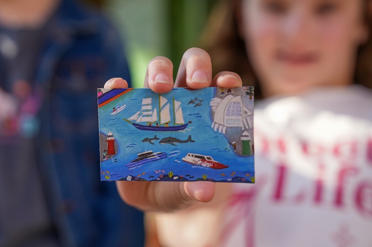 A child's hand holding up a small puzzle piece with an illustration of a seaside scene on it.
