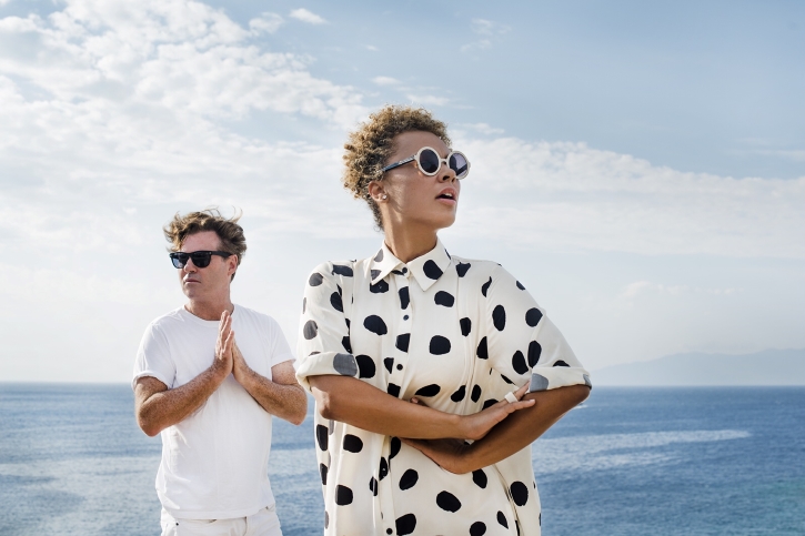 A woman in a black and white polka dot shirt and man in a white shirt stand in front of a windswept ocean.