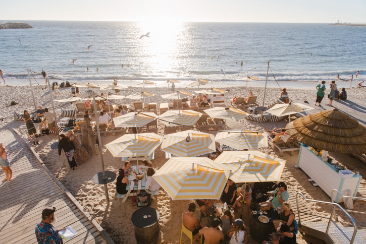 A group of yellow striped beach umbrellas with people underneath them on a beach watching the sunset.