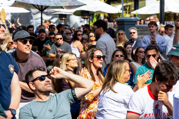 A crowd of people watching sport in a beer garden on a sunny day.