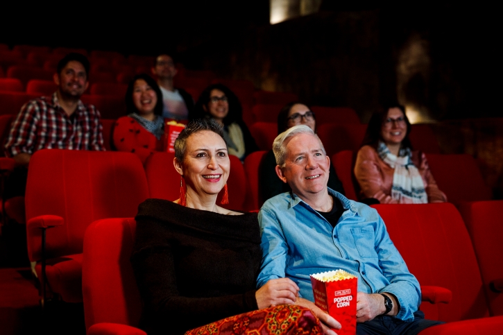 A man in a blue shirt and woman in a black top sitting in a movie theatre sharing some popcorn.