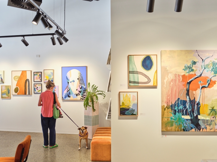 A woman with a small dog on a lead perusing art in a gallery.