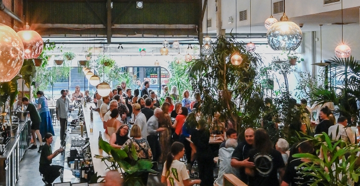 A large bar and dining room with lots of greenery and colourful lights hanging from the ceiling, with lots of people socialising below.