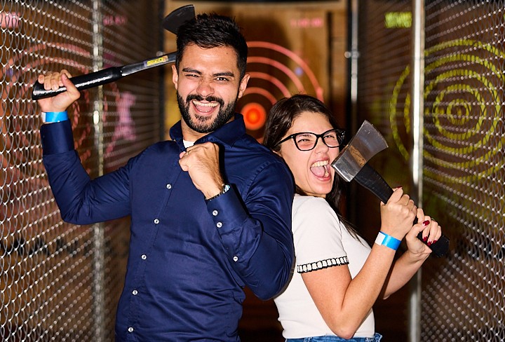 A young man and young woman with their backs to each other wielding axes and pulling funny faces.