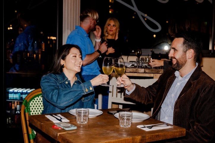 A man and a woman dining in a dimly let restaurant, clinking their glasses together.