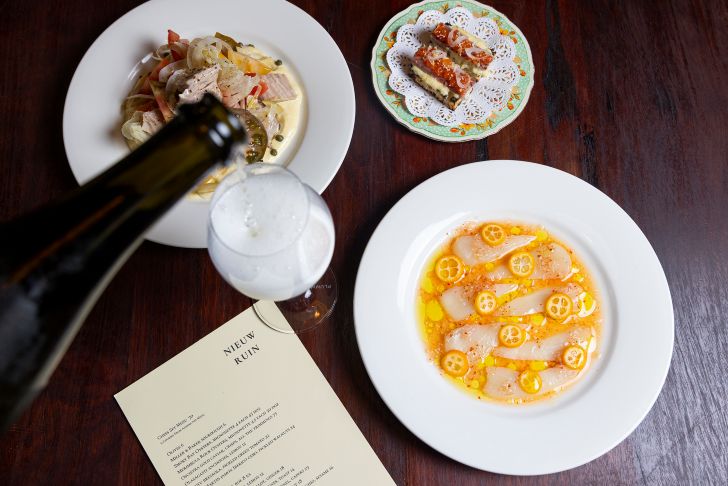 A trio of dishes on a dark wooden table with a glass of champagne being poured.