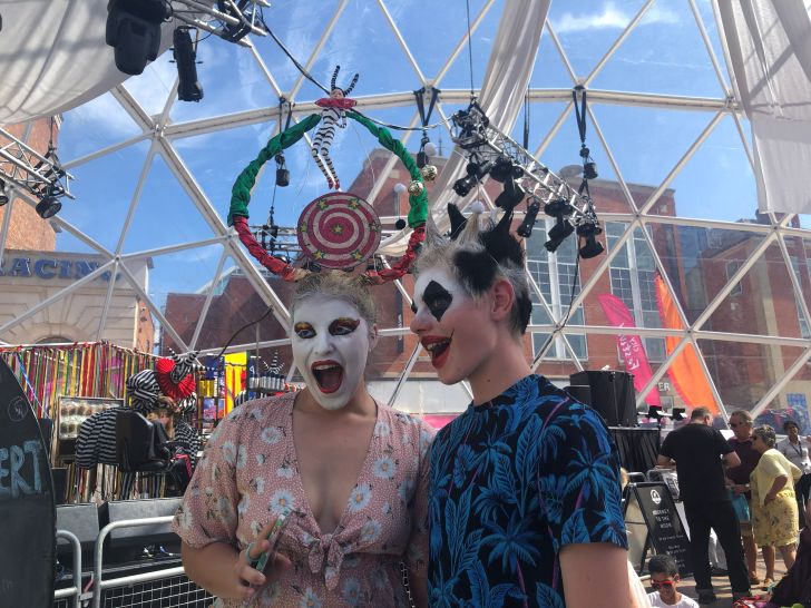 Two young women wearing black and white face paint and extravagant, colourful hairstyles with ornaments hanging from their hair.