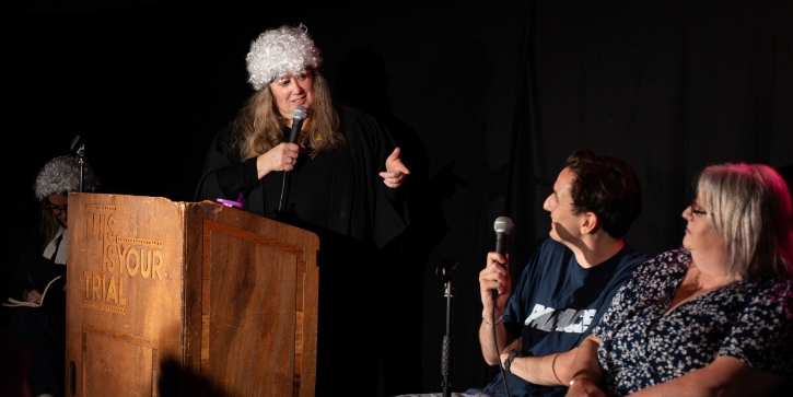 A woman in a barrister's wig stands behind a podium pointing at a man and woman sitting by her.