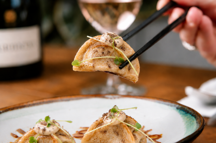 a dumpling being lifted by chopsticks with a glass of wine in the background