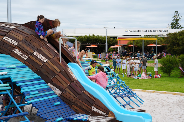 kids sliding down a dolphin installation with a blue slide and people in the background outside a cafe with an orange cafe in the backgroun