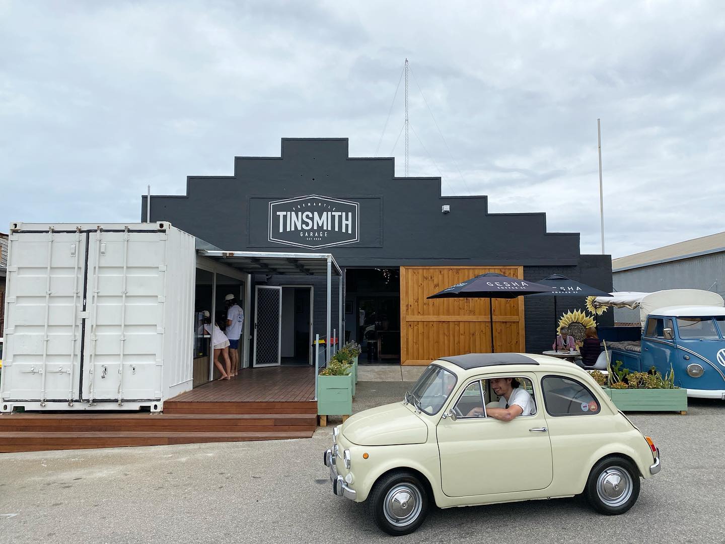 a smiling man in a vw outside a cafe called tinsmith with a few people lined up at a window to order