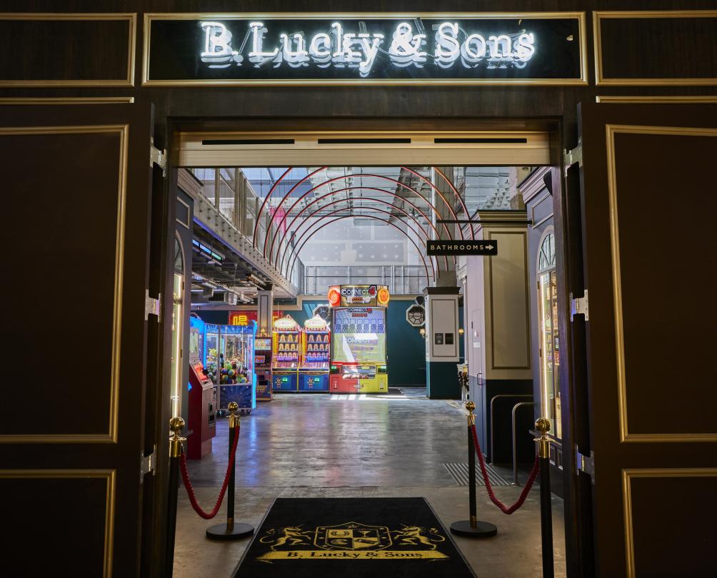 Entryway to B. Lucky & Sons arcade with dark wooden doors, rope bollards and bright neon lights of arcade games visible in the distance 