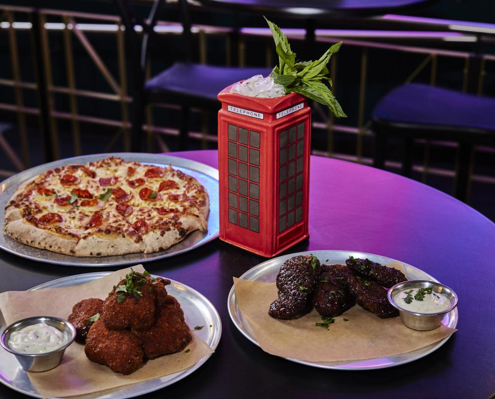 Pizza and other snacks plated up and presented on a table next to a cocktail served in a London-style red telephone booth
