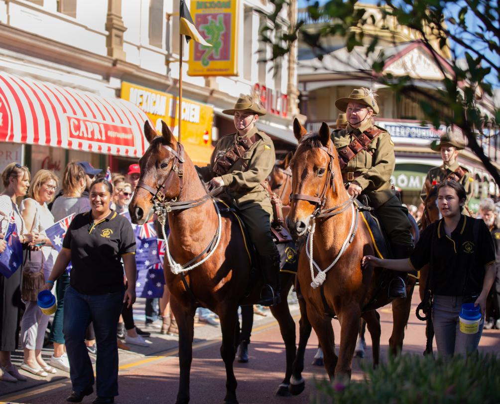 service people in uniform on horses walking through crowded street 