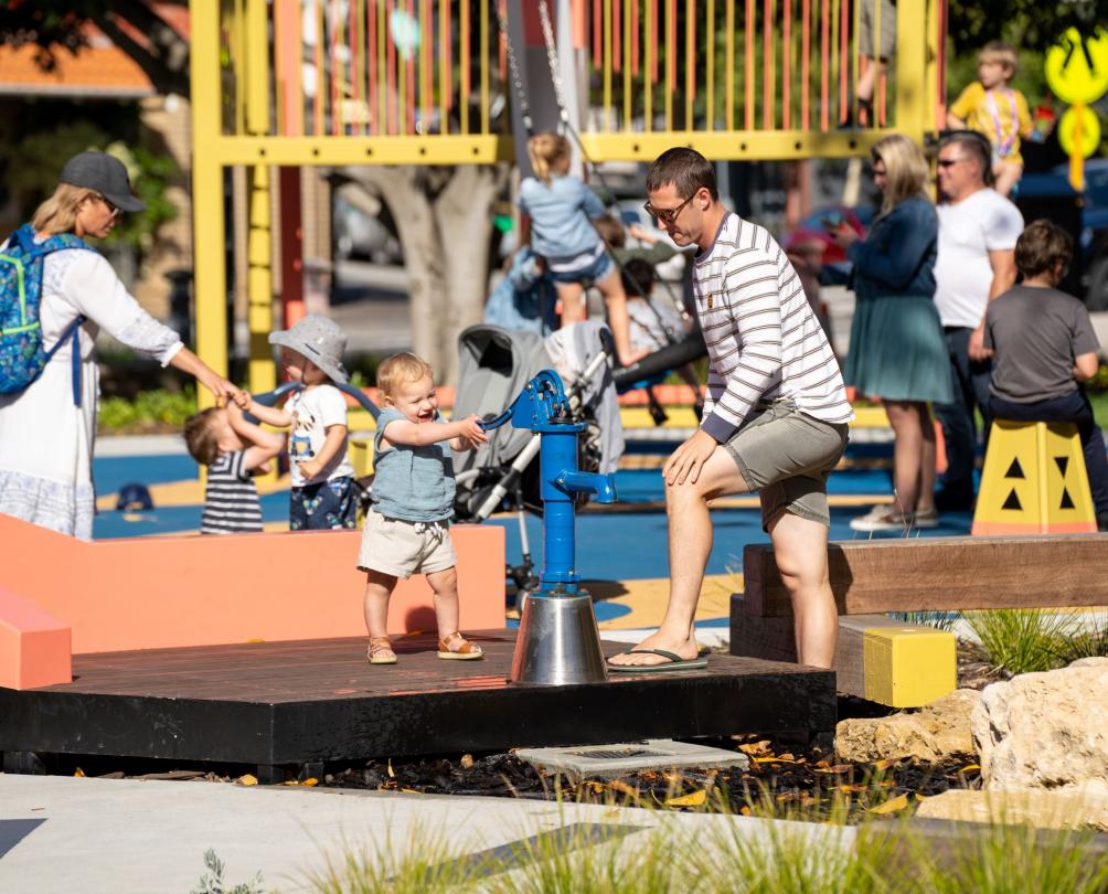 Federal Hotel Child playing with water pump, watched by parent at Walyalup Koort playground