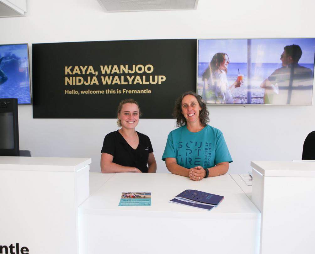 two women stand behind counter smiling. Black sign in the background with text Kaya, Wanjoo Nidja Walyalup 