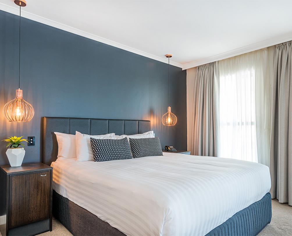 interior of hotel room with made up bed and two bedside tables with lights in front of a blue wall