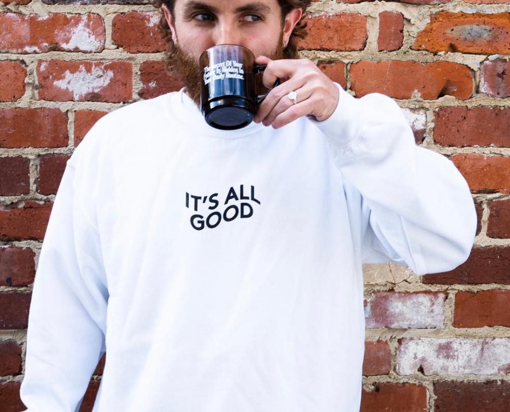 Man in white jumper that reads 'It's All Good' drinking a coffee from a mug