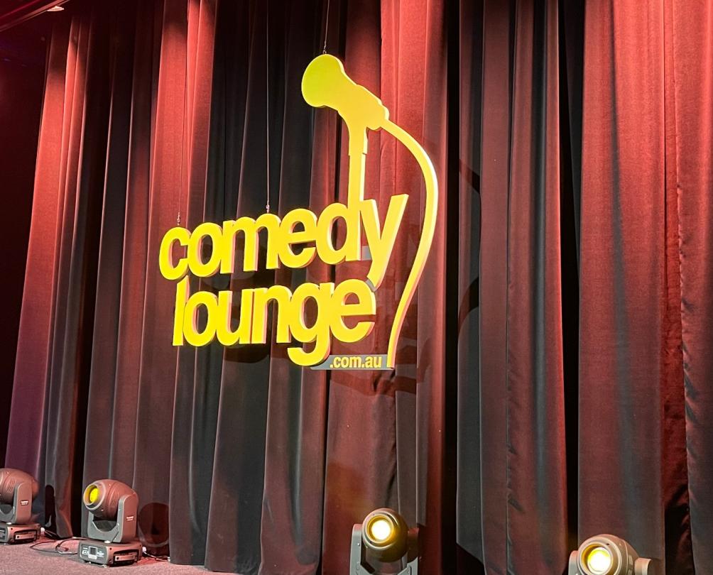 A red stage curtain with yellow neon sign 'Comedy Lounge' upstairs at Sailing for Oranges 