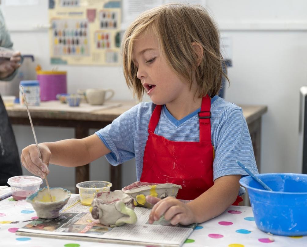 Young child in red apron painting clay works at Fremantle Arts Centre kids courses