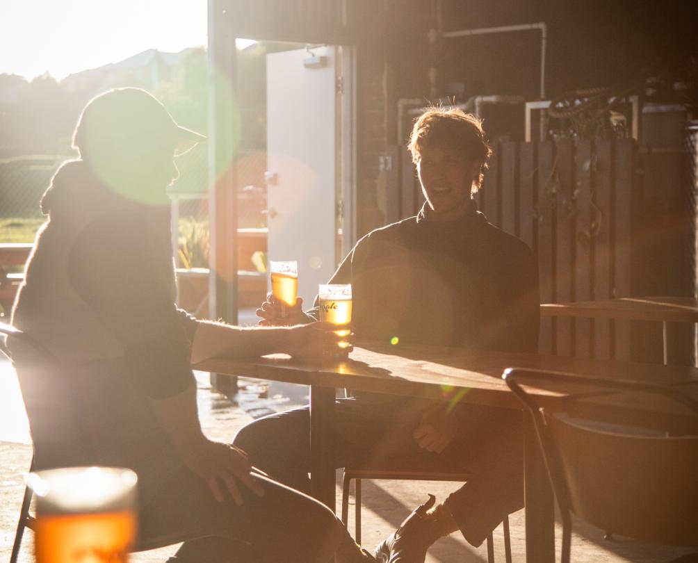 Two men sitting at a small table in a shed drinking a couple of beers.