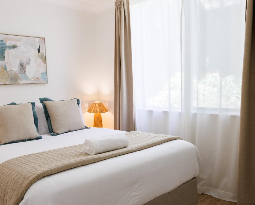 A white bed with linen sheets and white curtains by a window.