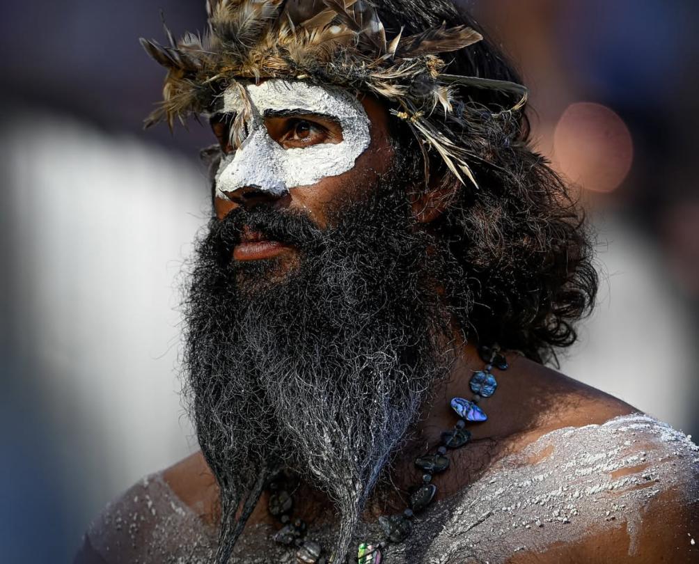 An Australian indigenous man with a long beard and white face and body paint wears a traditional feathered headpiece.