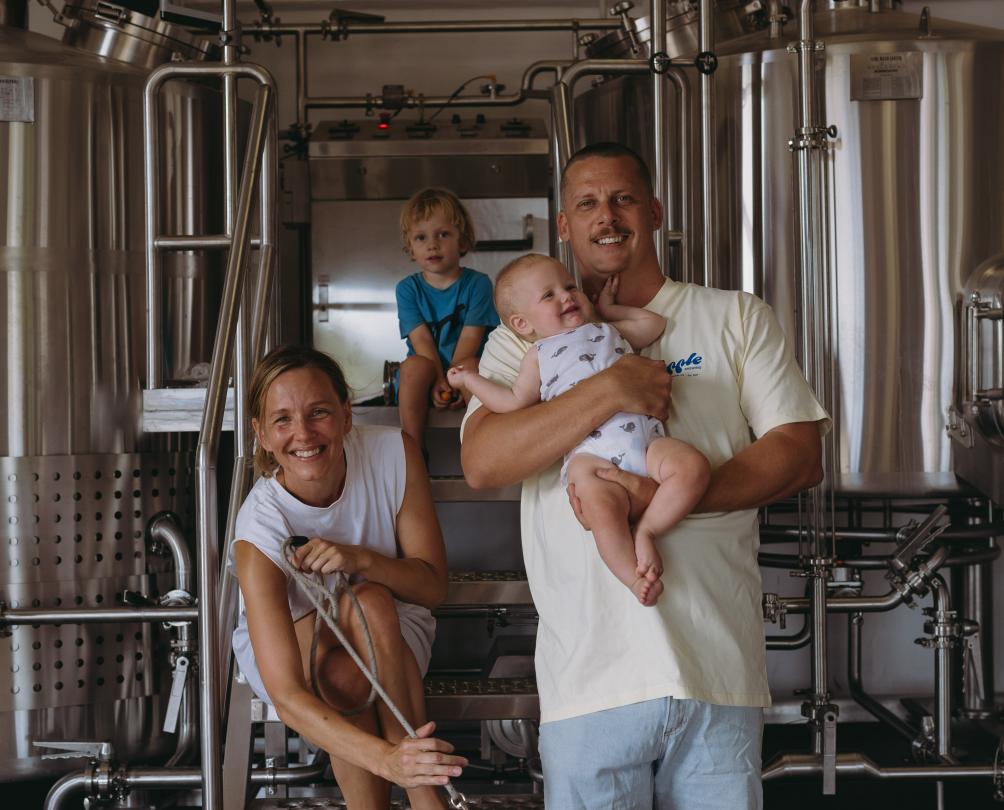 A couple with two young kids and a greyhound standing in front of brewing equipment.