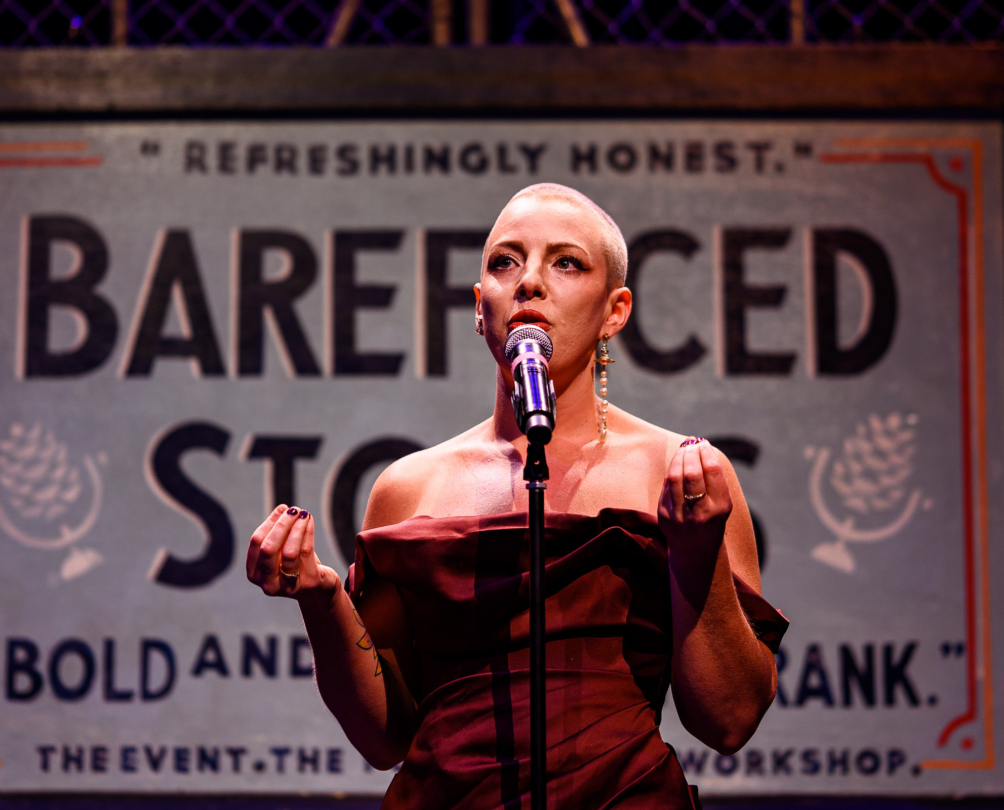 A woman speaks into a microphone on a stage with the words 'Barefaced Stories' behind her.