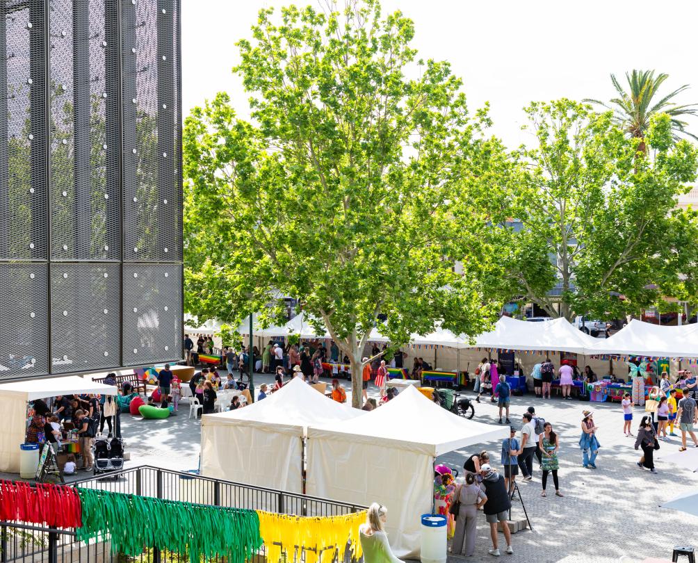 A city square with several stalls and marquees milling with people underneath several tall trees.