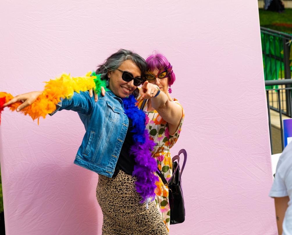 Two people pose in front of a pink photo wall wearing rainbow scarves.