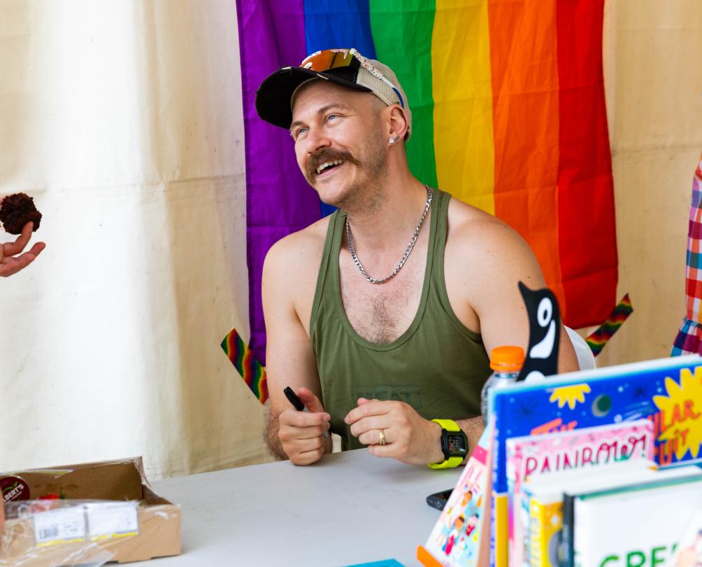 A person sitting at a table in front of a rainbow flag talking to someone.
