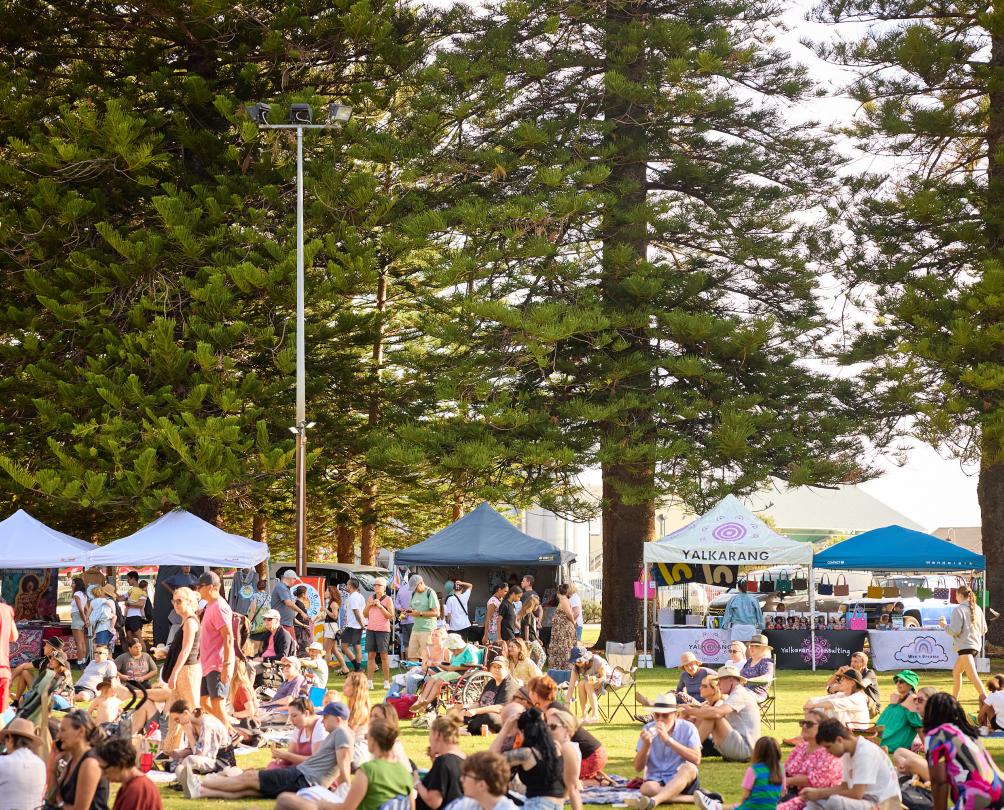 A crowd of people with picnic blankets and chairs sit on lawn with food stalls behind them.
