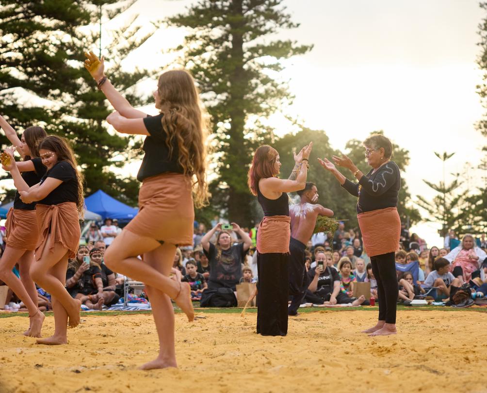 A group of young Aboriginal women perform a traditional dance on sand.