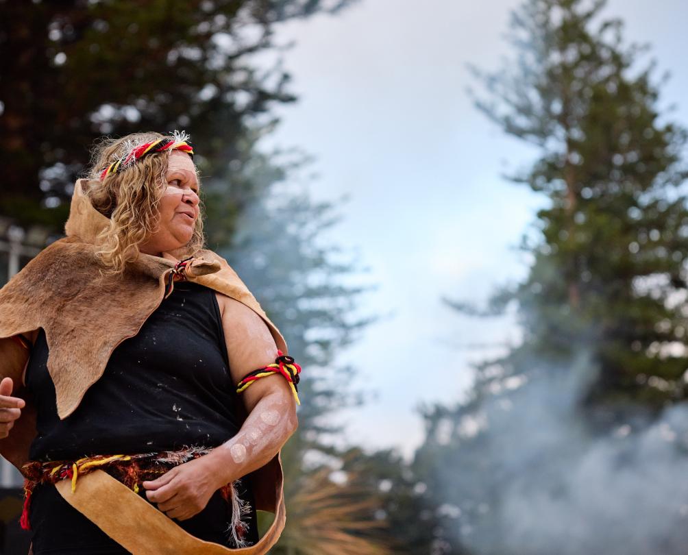 An indigenous woman in traditional dress and body paint with smoke and tall green pines behind her.