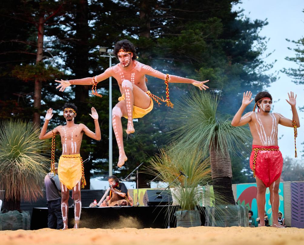Three young Aboriginal men perform a traditional dance, one leaping high into the air.