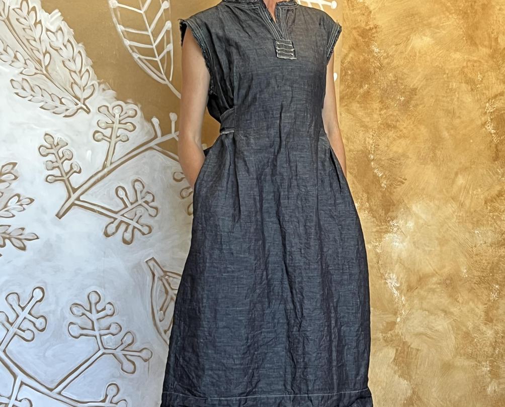 A woman in a grey dress stands against a yellow patterned wall.