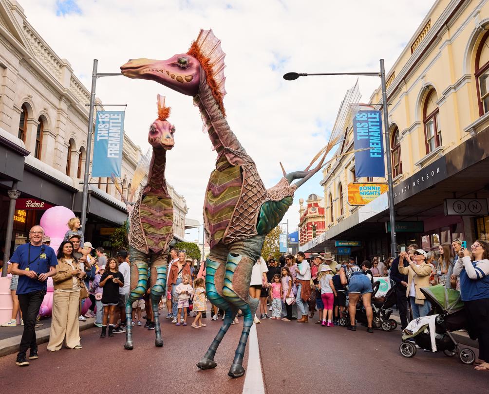 Two people on stilts wearing elaborate fantasy costumes walk down a busy street.