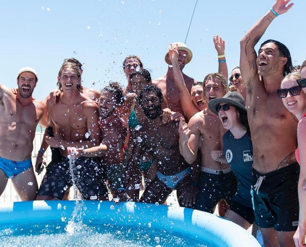 A group of people cheering around a large inflatable pool on a beach.