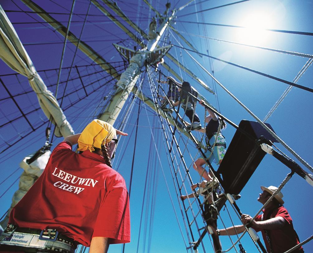 A crew member of a tall ship watches as three other crew members climb a mast.